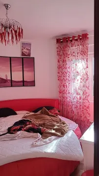 Spavaća soba sa neurednim krevetom, crvenim zavesama i slikama na zidu. - image 11