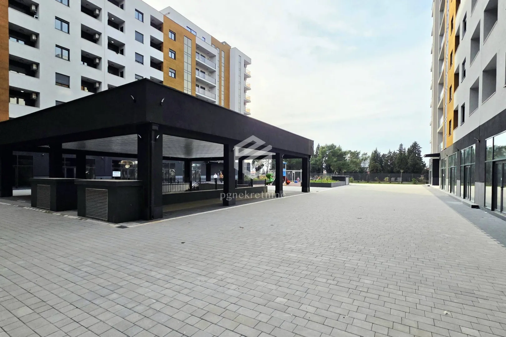 Pogled na popločano dvorište i moderne stambene zgrade sa nadstrešnicom.