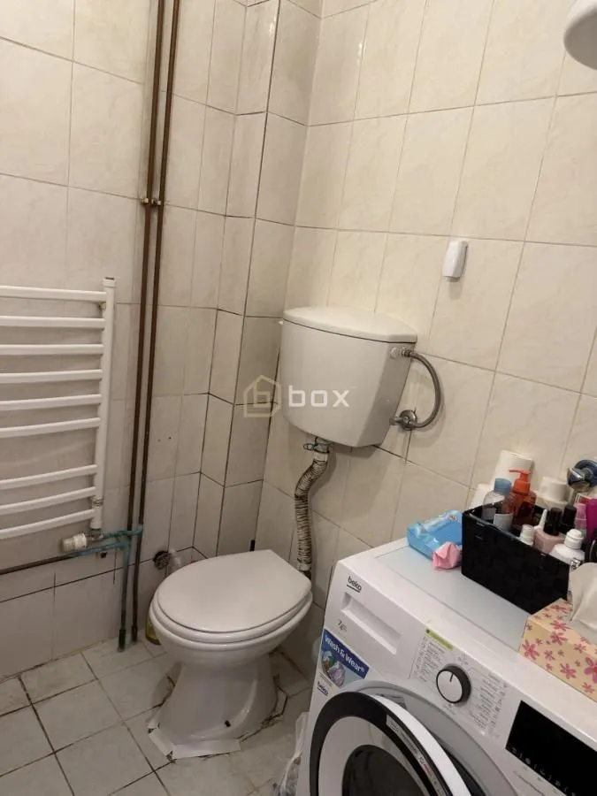 Manje kupatilo sa toaletom i veš mašinom obloženo svetlim pločicama.