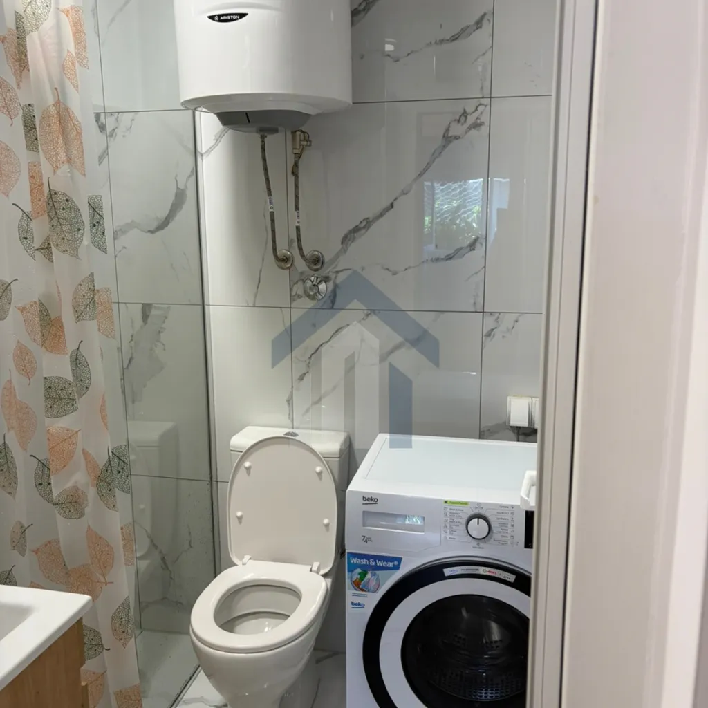 Manje kupatilo sa bijelim pločicama, toaletom i veš mašinom.