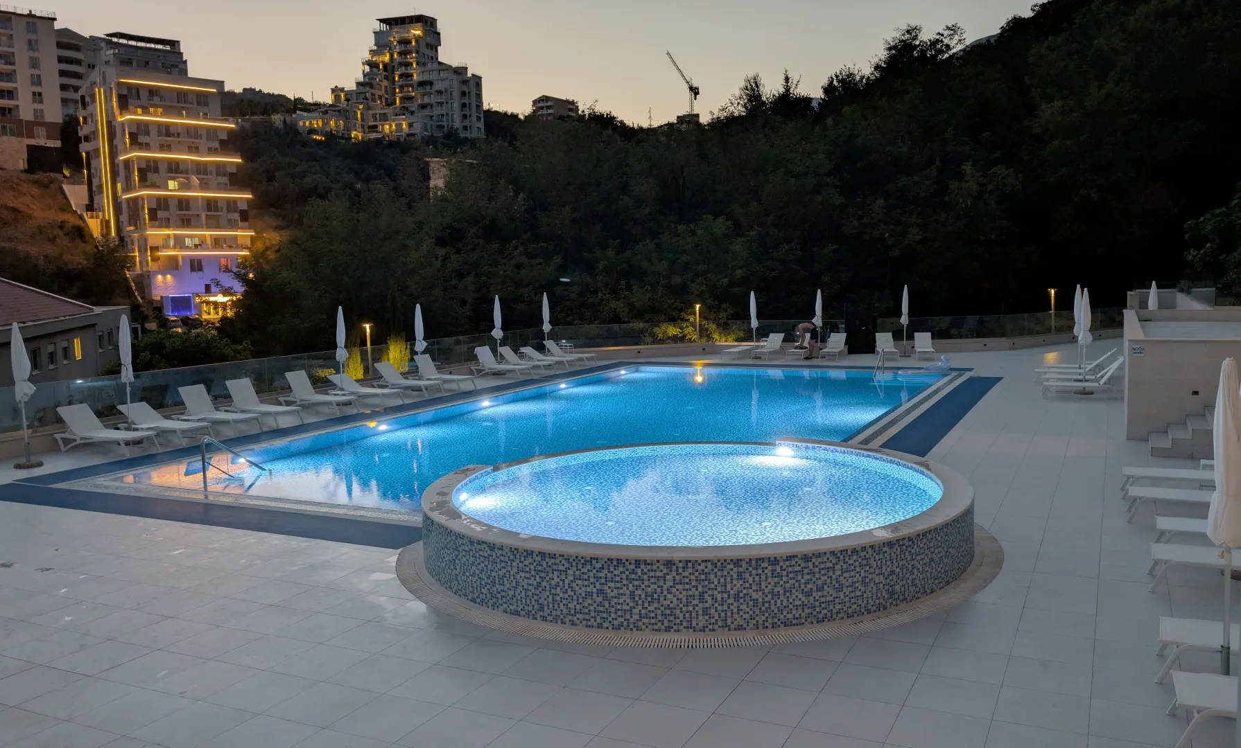 Modern swimming pool with loungers and a view of surrounding buildings and hills at dusk.