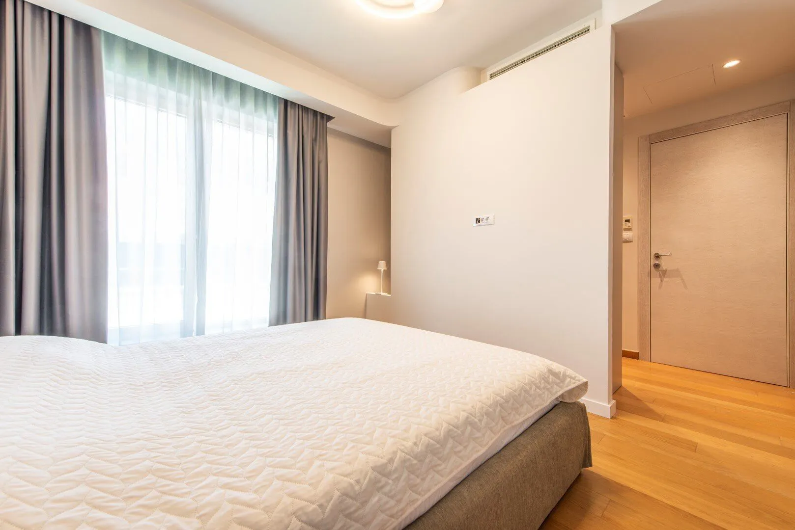 Svjetla spavaća soba sa bračnim krevetom, drvenim podom i prozorom sa zavjesama.