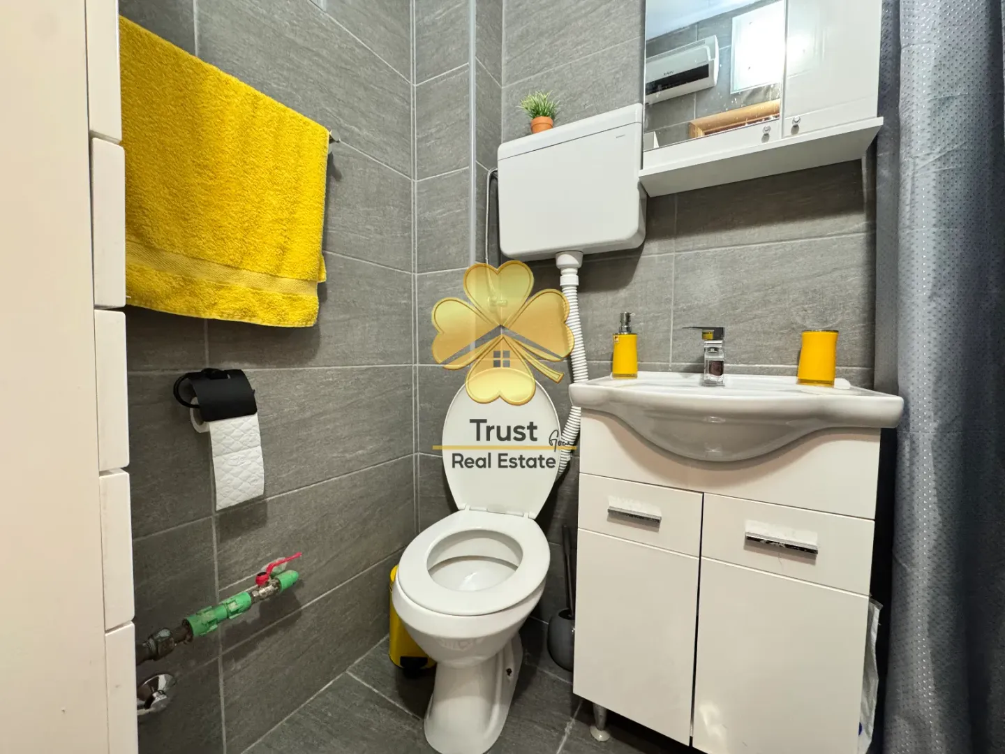 Manje kupatilo sa sijem, lavaboom i wc šoljom obloženo sivim pločicama.