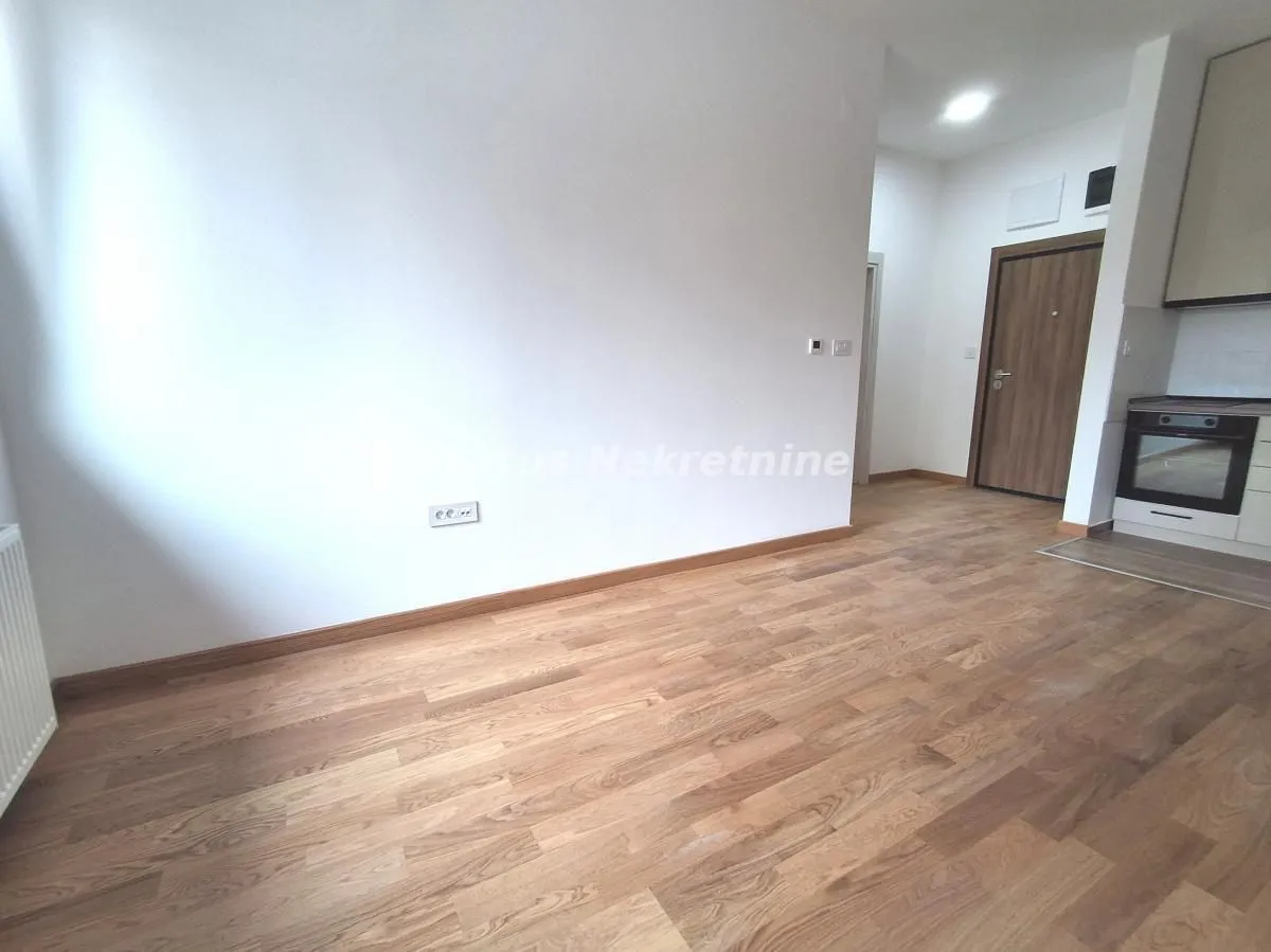 Empty room with wooden flooring and a partial view of the kitchen in the corner.