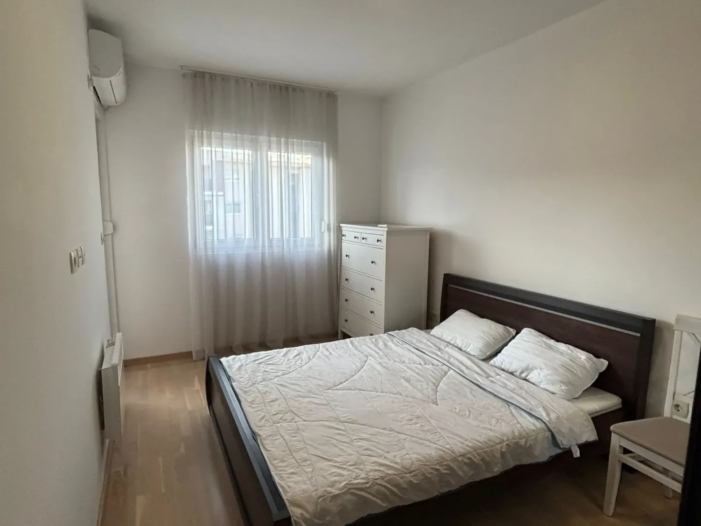 Furnished bedroom with a double bed, white chest of drawers, and a window with curtains.