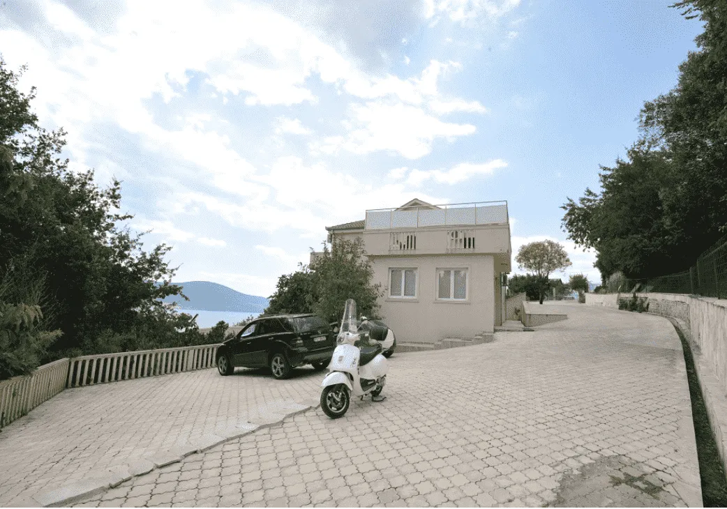 Pogled na spoljašnjost kuće sa dvorištem, parkingom i parkiranim vozilima.