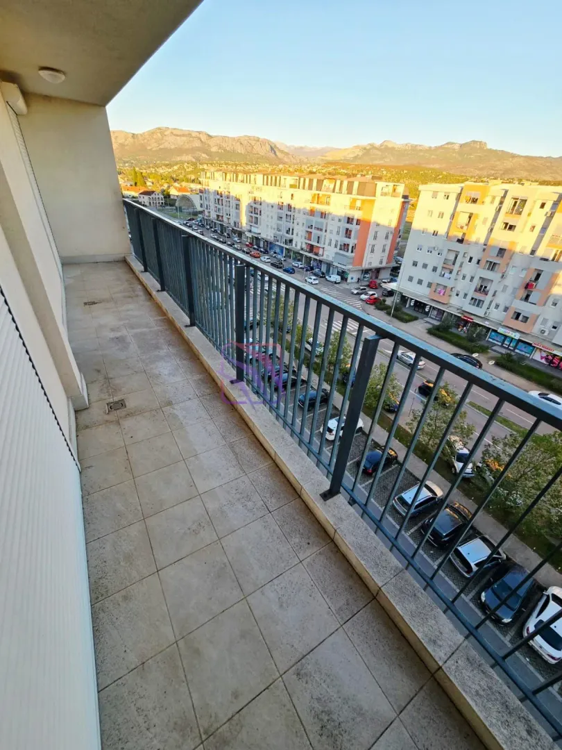 Dugi popločani balkon sa metalnom ogradom i pogledom na grad i planine.