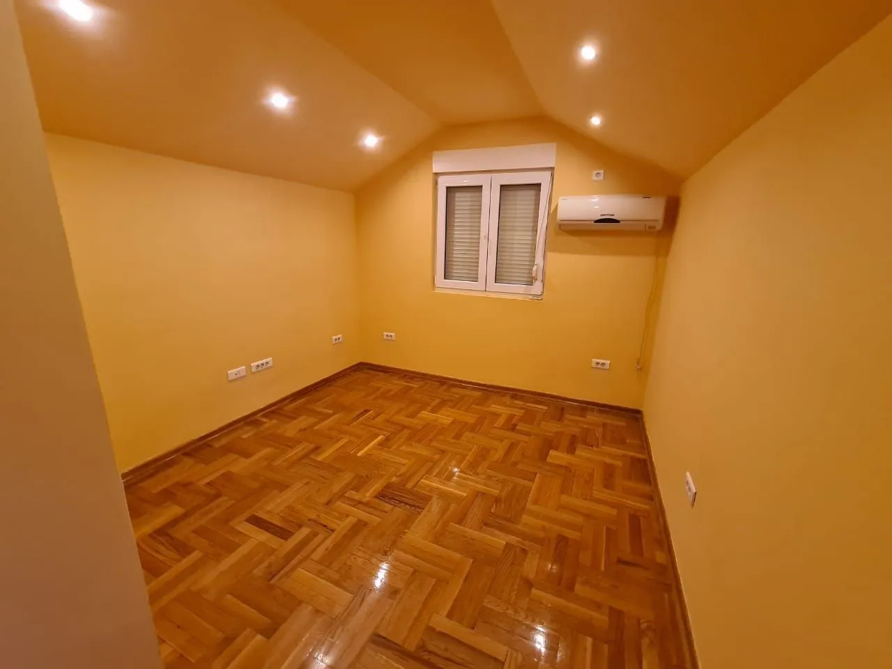 Prazna soba sa žutim zidovima, drvenim parketom i klima uređajem.