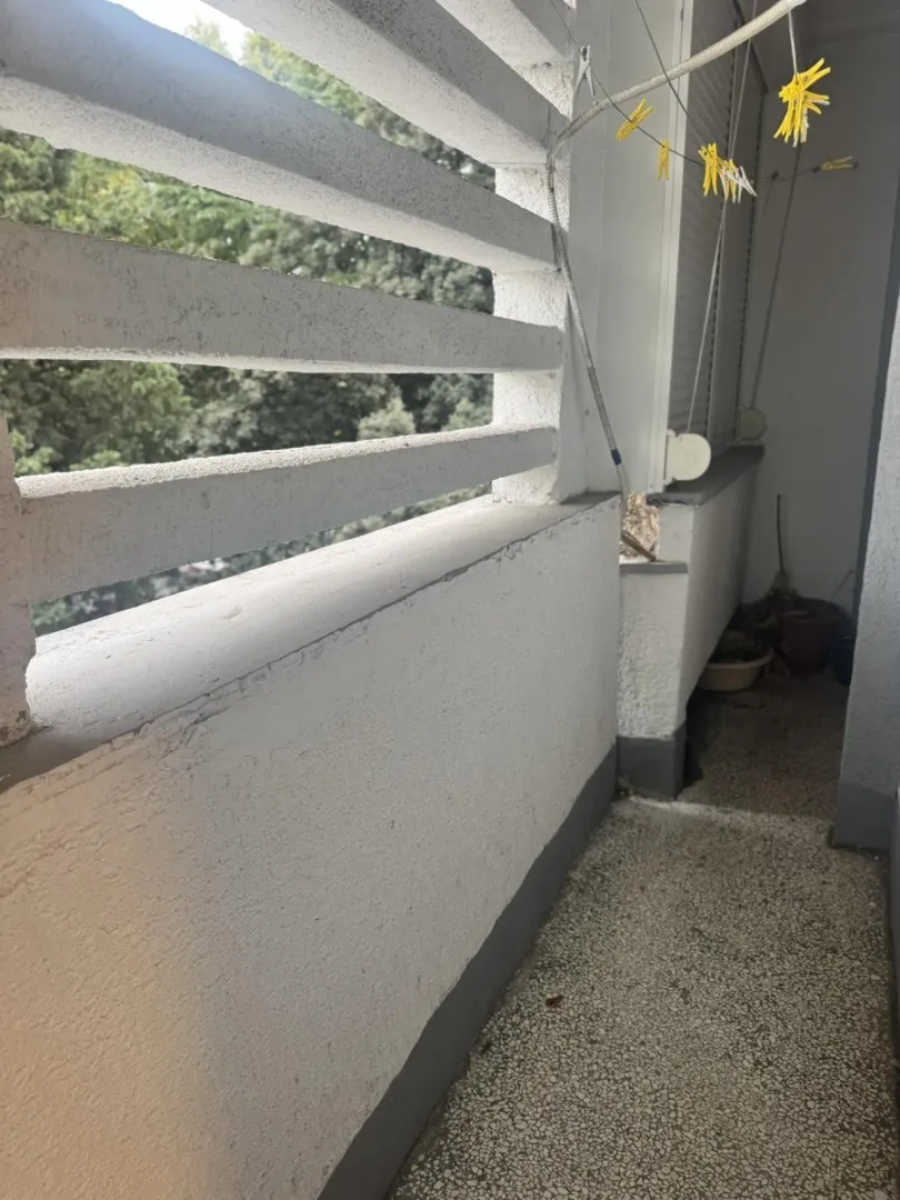 Uski balkon sa betonskom ogradom i pogledom na drveće.