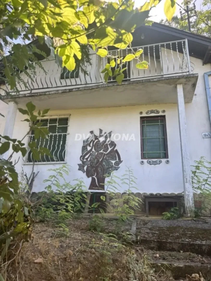 Pogled na fasadu dvospratne kuće sa balkonom i zelenilom u dvorištu.