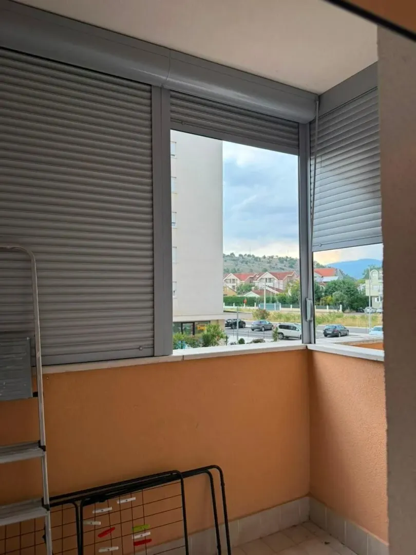 Natkriveni balkon sa roletnama i pogledom na okolne zgrade i brda.