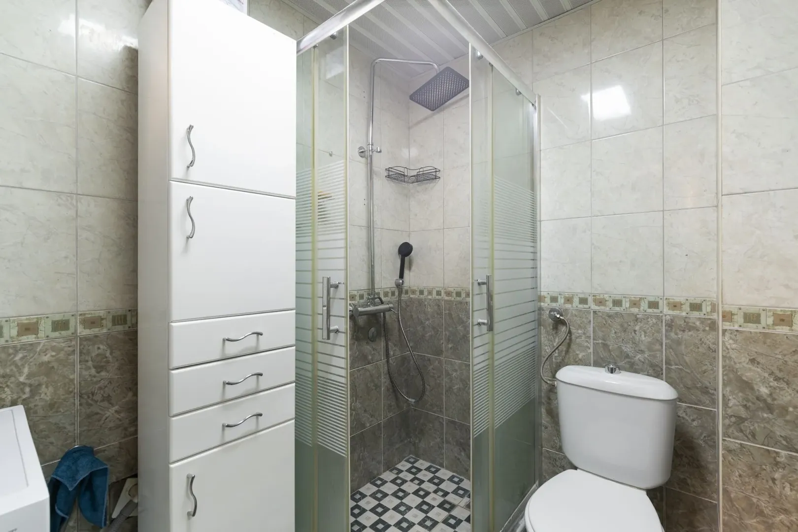 Kupatilo sa tuš kabinom, WC šoljom i bijelim ormarićem.