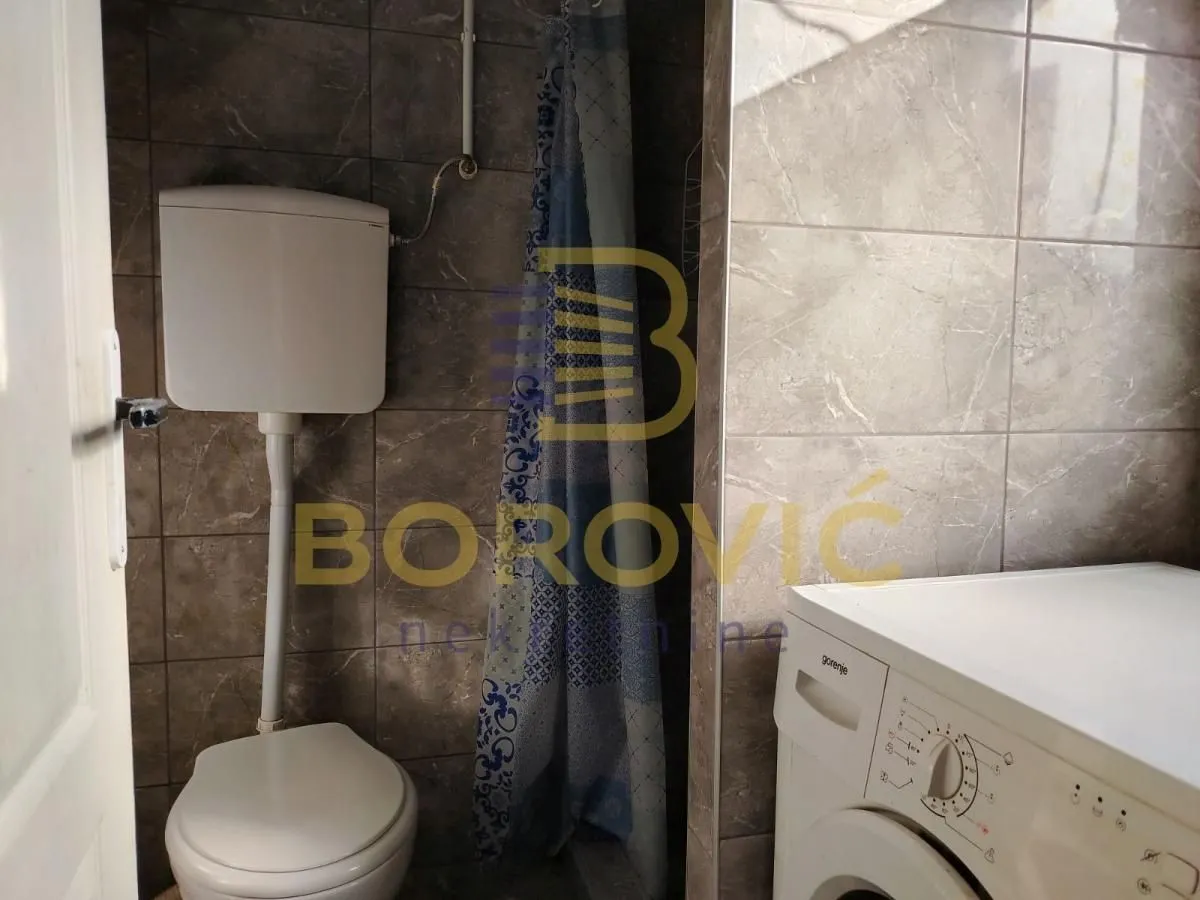 Manje kupatilo sa wc šoljom, tuš kabinom i veš mašinom obloženo tamnim keramičkim pločicama.