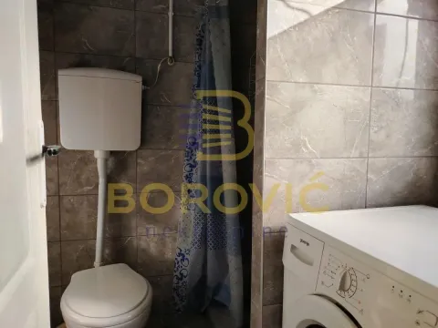Manje kupatilo sa wc šoljom, tuš kabinom i veš mašinom obloženo tamnim keramičkim pločicama. - image 3
