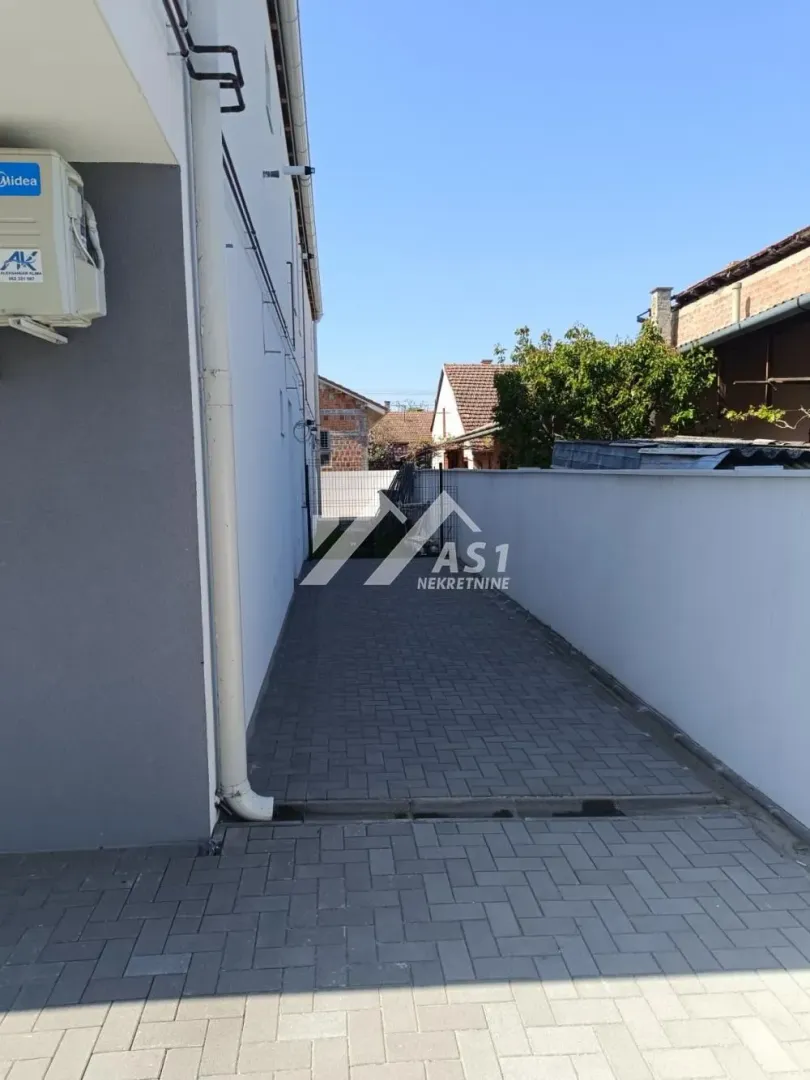 Popločani prolaz pored kuće sa ogradom.