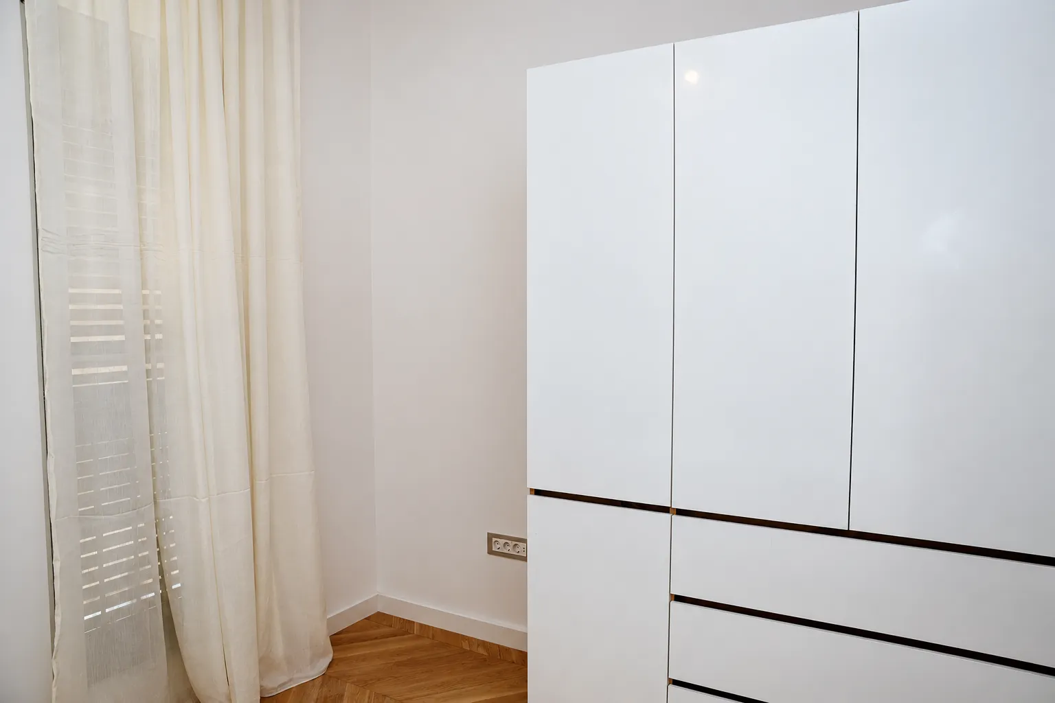 Bijeli garderober u sobi sa drvenim podom i prozorom sa zavjesama.