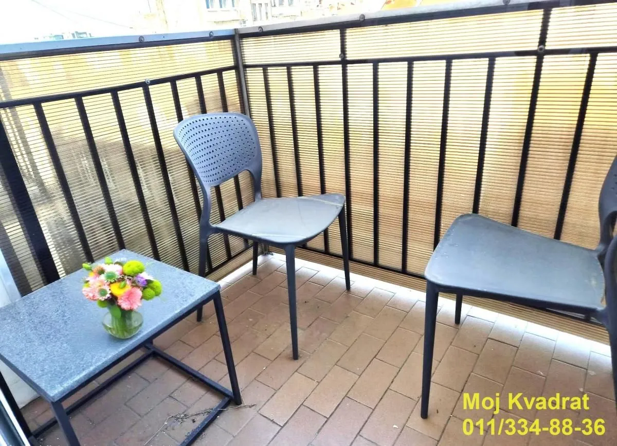 Manji balkon sa crnom ogradom, stolom i dve stolice.
