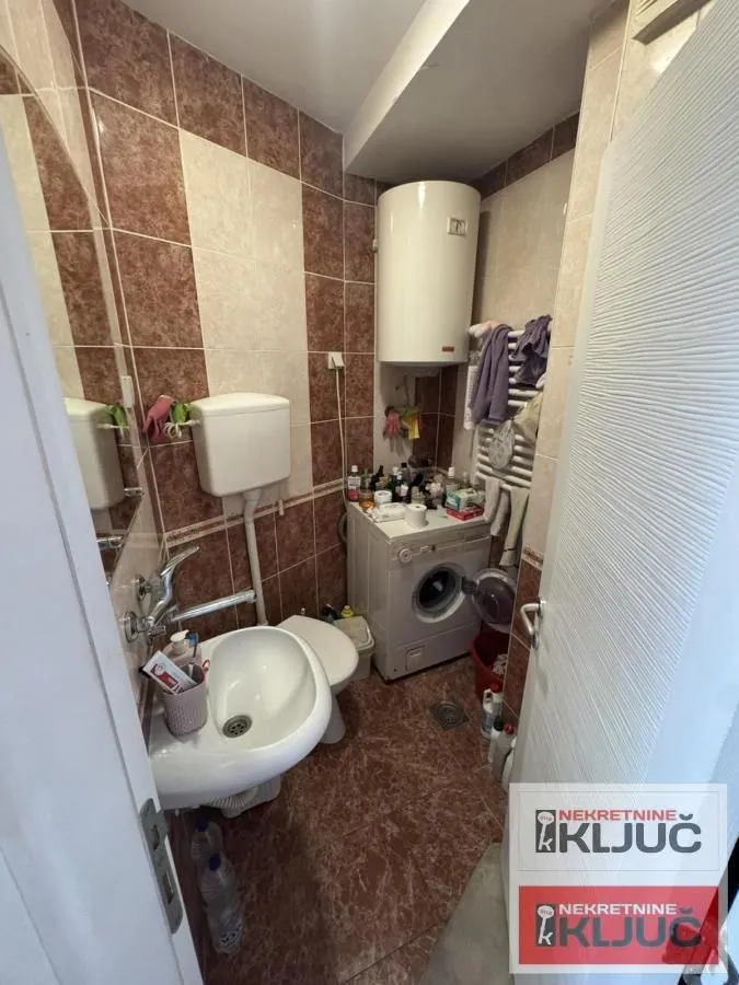 Manje kupatilo obloženo keramičkim pločicama sa tuš kabinom, umivaonikom i veš mašinom.