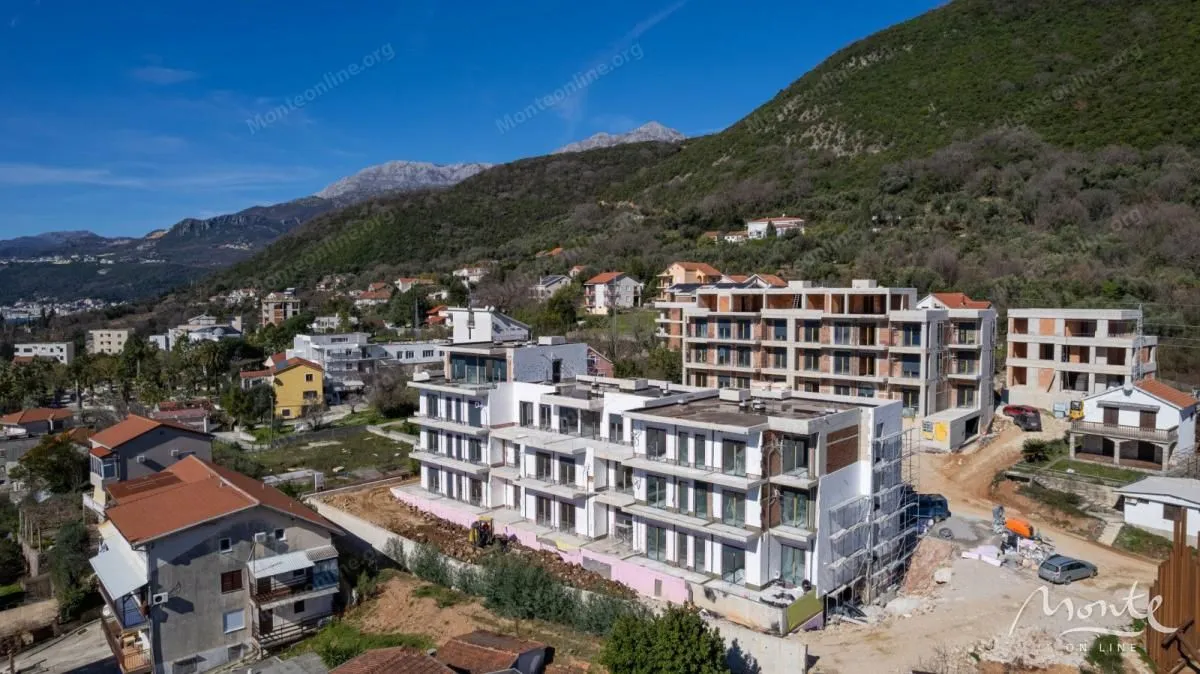 Pogled iz vazduha na stambene zgrade u izgradnji okružene zelenilom i planinskim pejzažom.