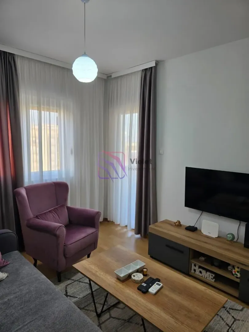 Dnevna soba sa garniturom, foteljom i TV komodom pored prozora sa zavesama.