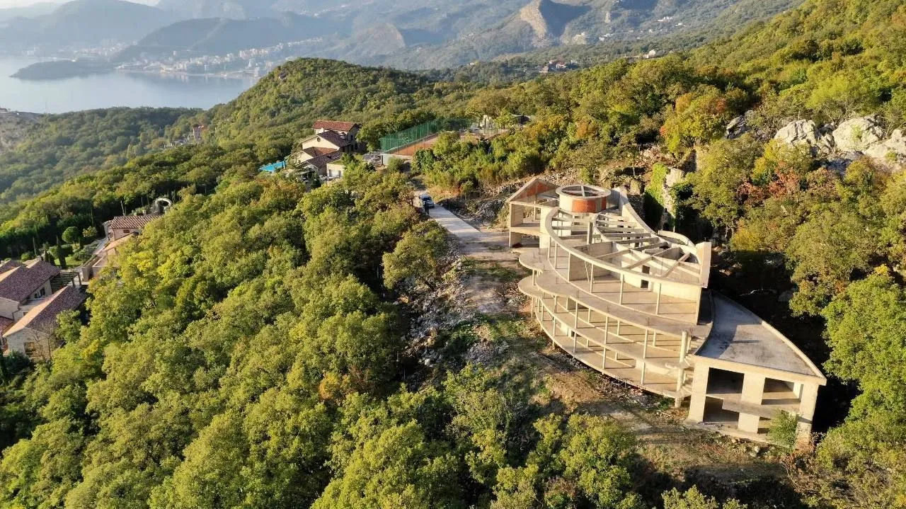 Pogled iz vazduha na kuću u izgradnji okruženu zelenilom i brdima.