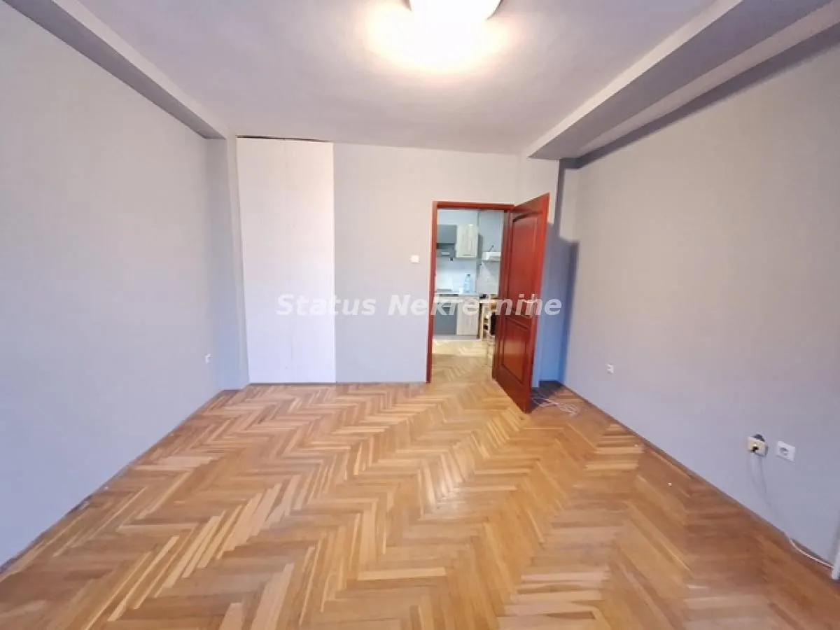 Prazna soba sa parketom i otvorenim vratima prema drugoj prostoriji.