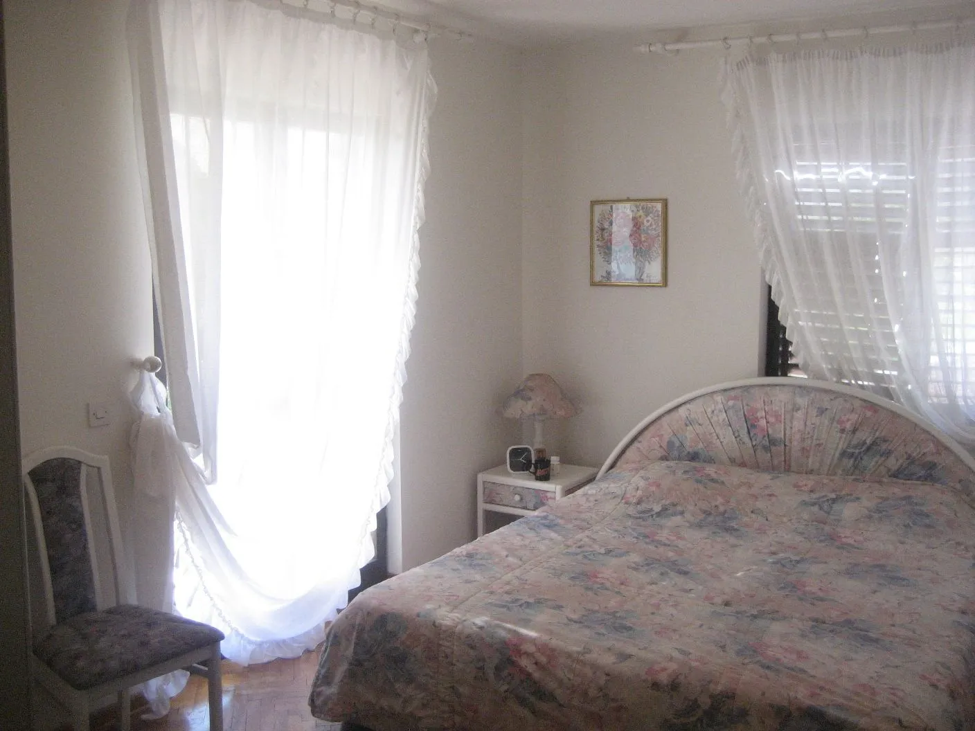 A bedroom featuring a double bed, a nightstand, and windows with white curtains.