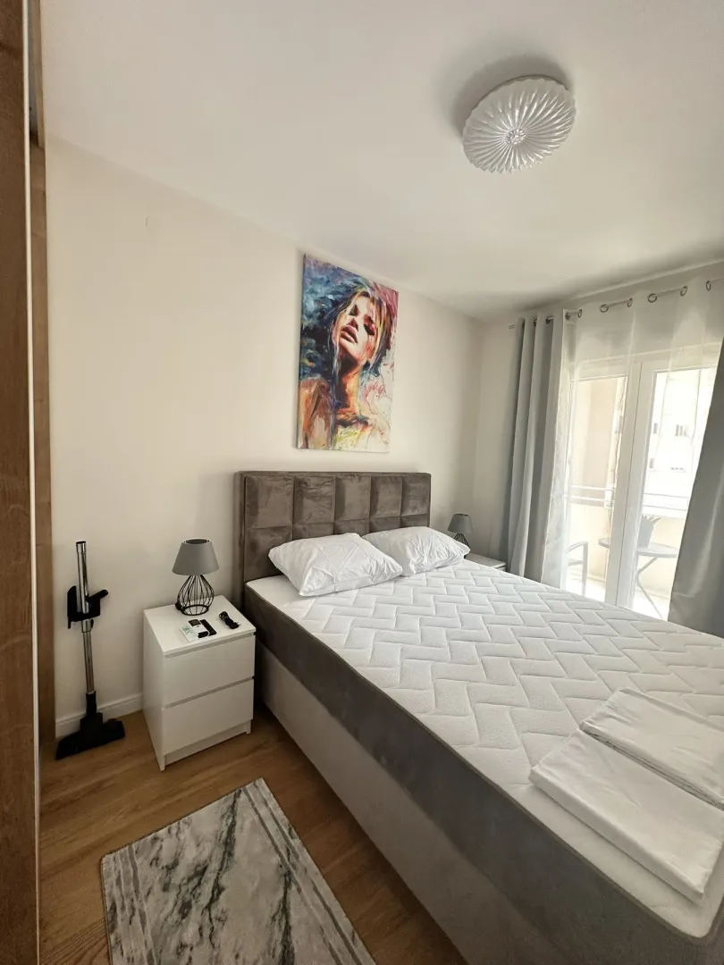 Svijetla spavaća soba sa francuskim ležajem, noćnim ormarićem i izlazom na balkon.