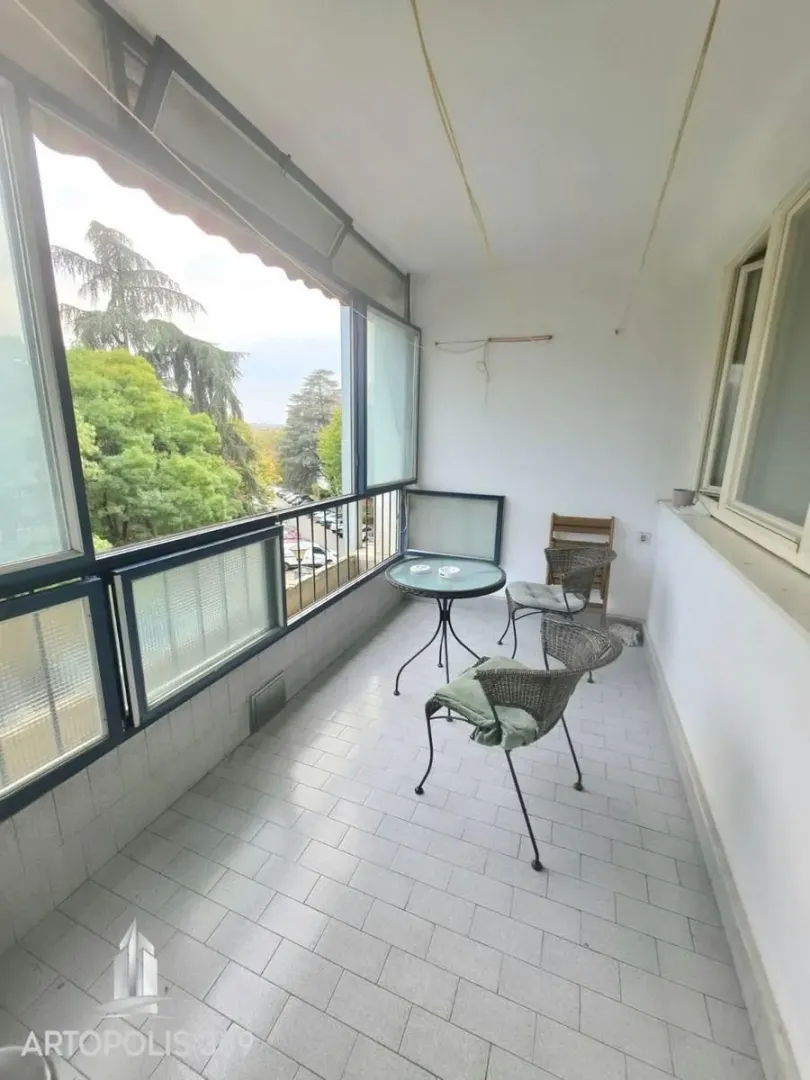 Zastakljeni balkon sa garniturom za sedenje i pogledom na zelenilo.