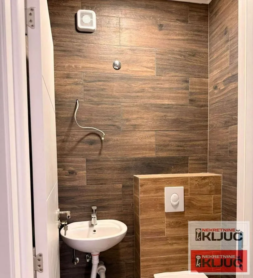 Manje kupatilo sa umivaonikom, WC šoljom i zidnim pločicama u dezenu drveta.