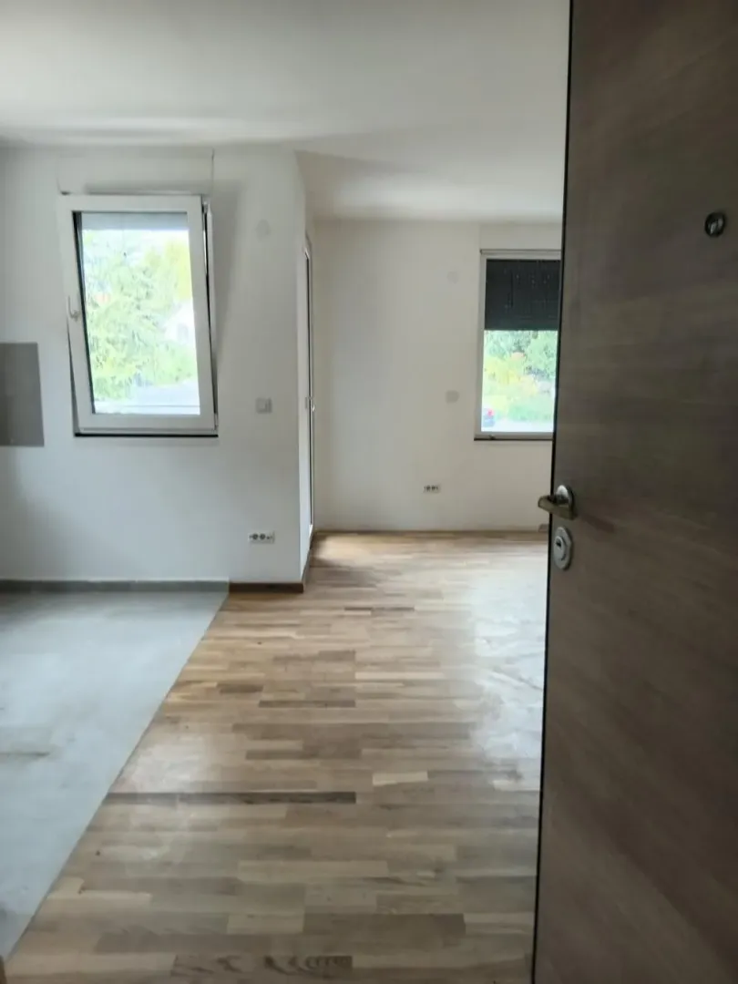 Pogled iz hodnika na nenamještenu prostoriju sa drvenim podom i prozorima.