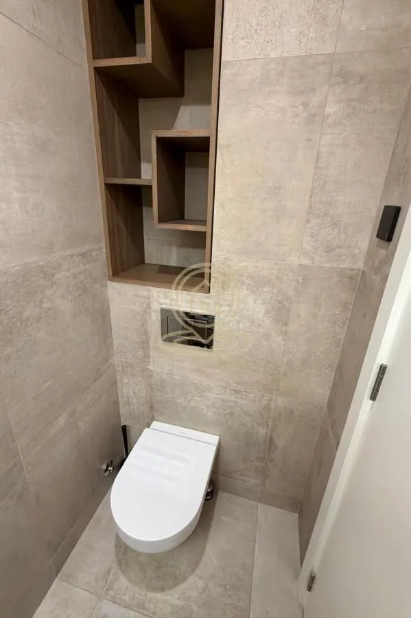 Manje kupatilo obloženo keramičkim pločicama sa ugradnom wc šoljom i drvenim policama.