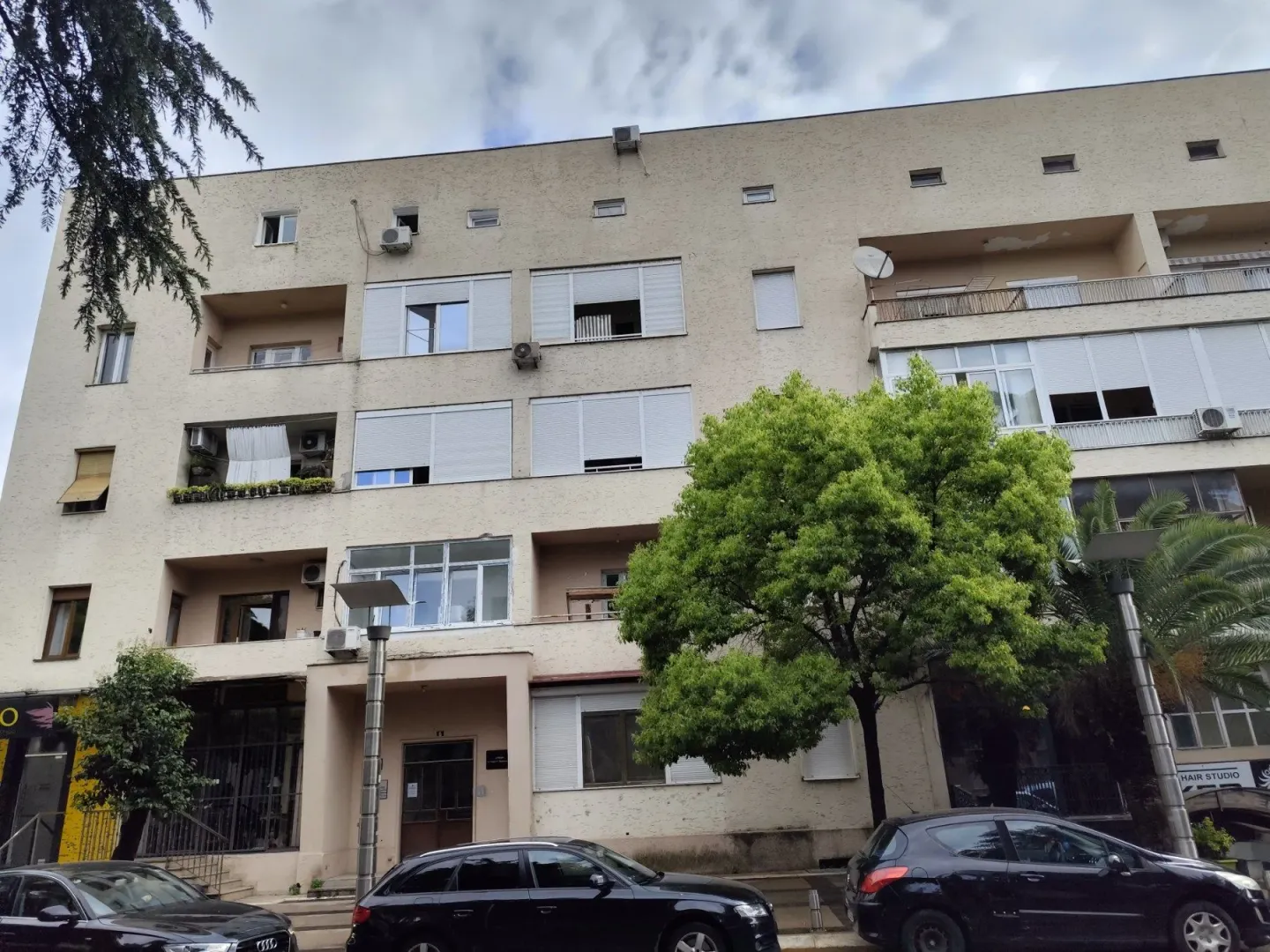 Spoljašnji izgled stambene zgrade sa balkonima i zelenilom ispred ulaza.