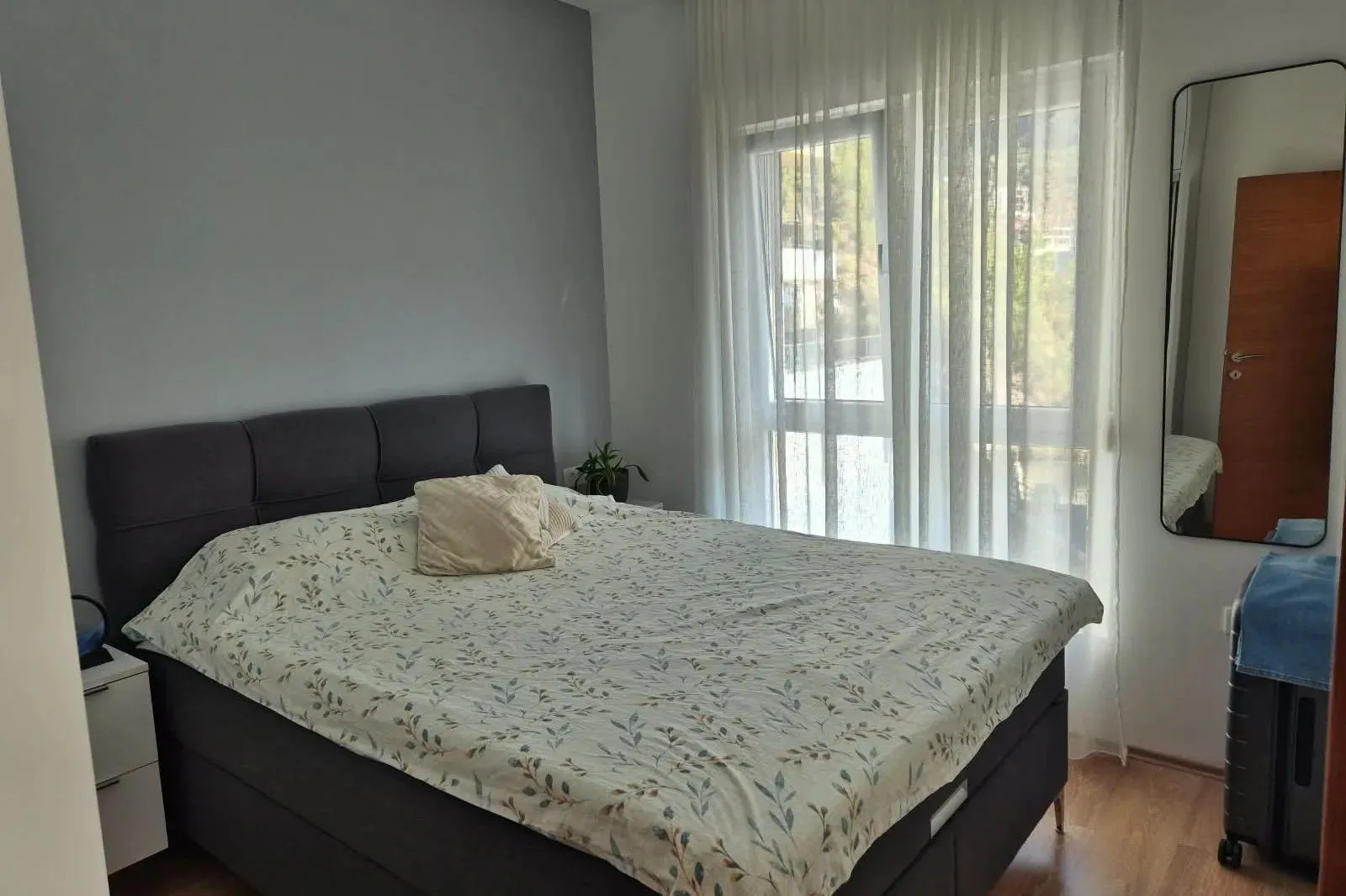 Furnished bedroom with a double bed, a window with curtains, and a mirror.