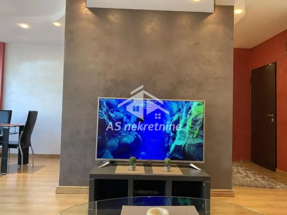 Dnevna soba sa televizorom na komodi i trpezarijskim stolom u pozadini.