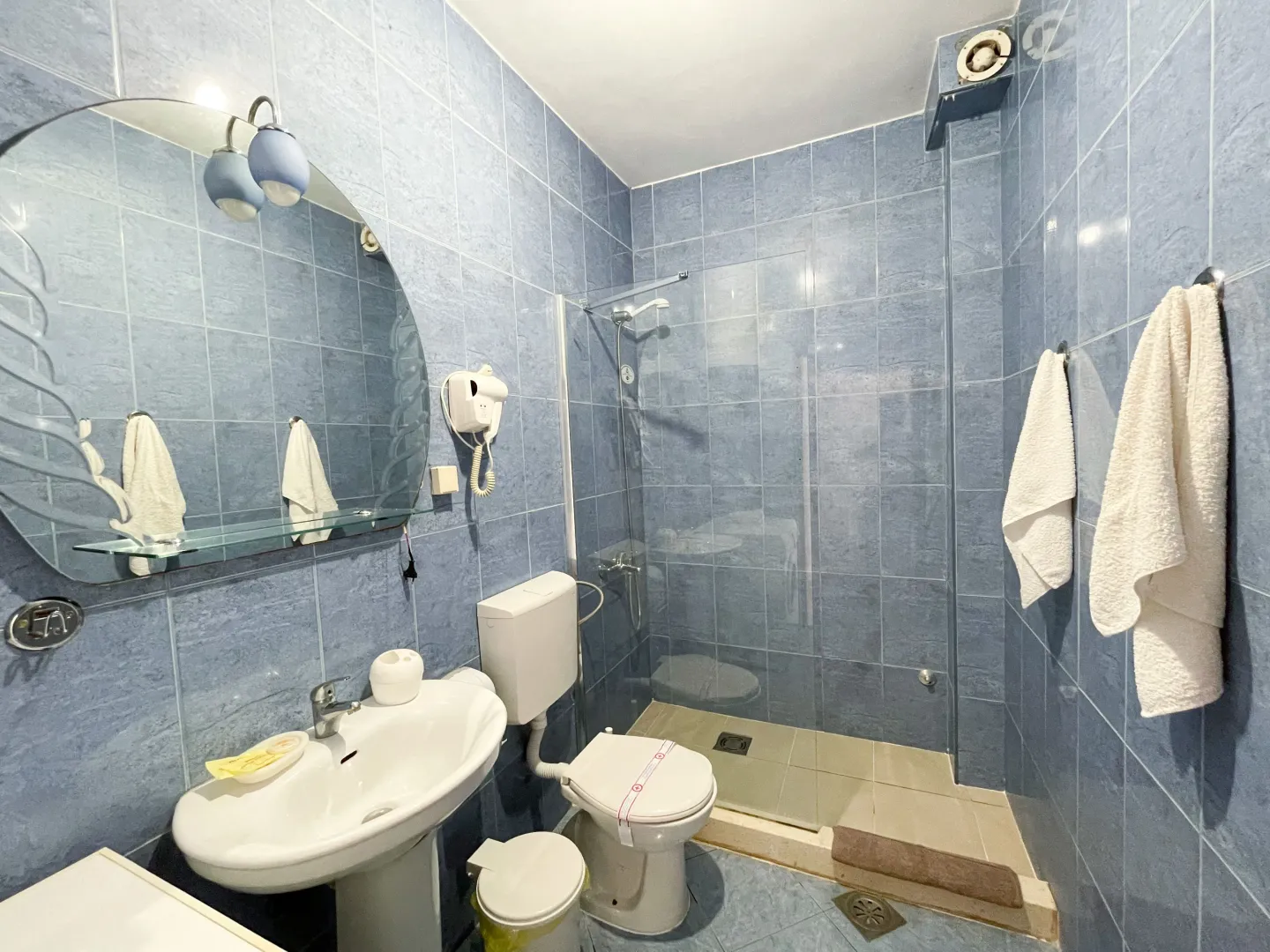 Kupatilo obloženo plavim pločicama sa umivaonikom, ogledalom i tuš kabinom.
