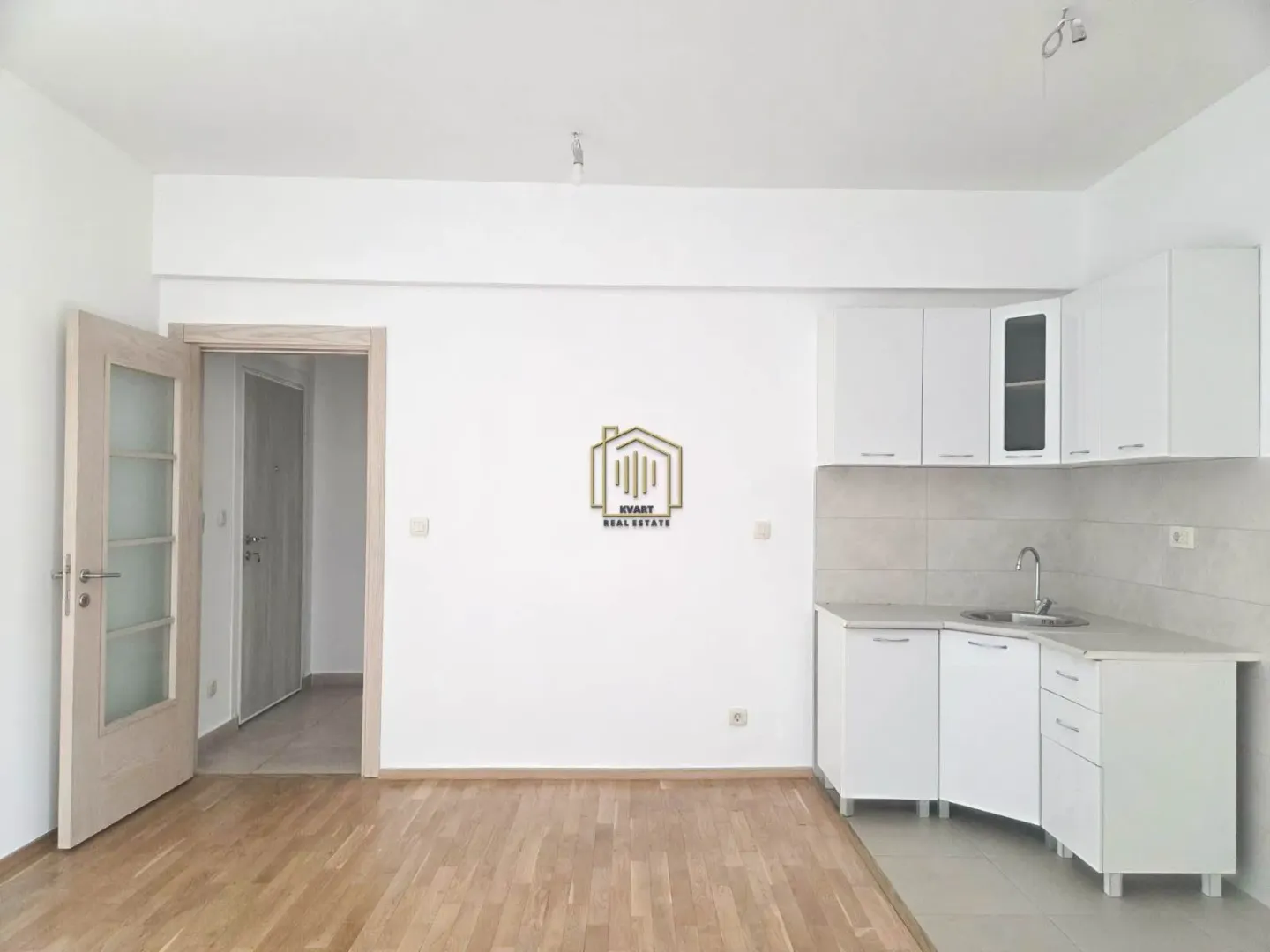 Manja bijela kuhinja u uglu prostorije sa drvenim podom.
