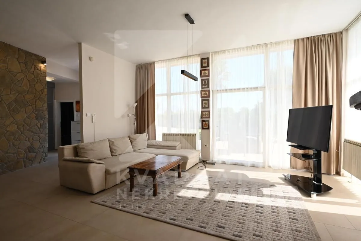 Spacious living room with a sofa set, large windows, and a TV.