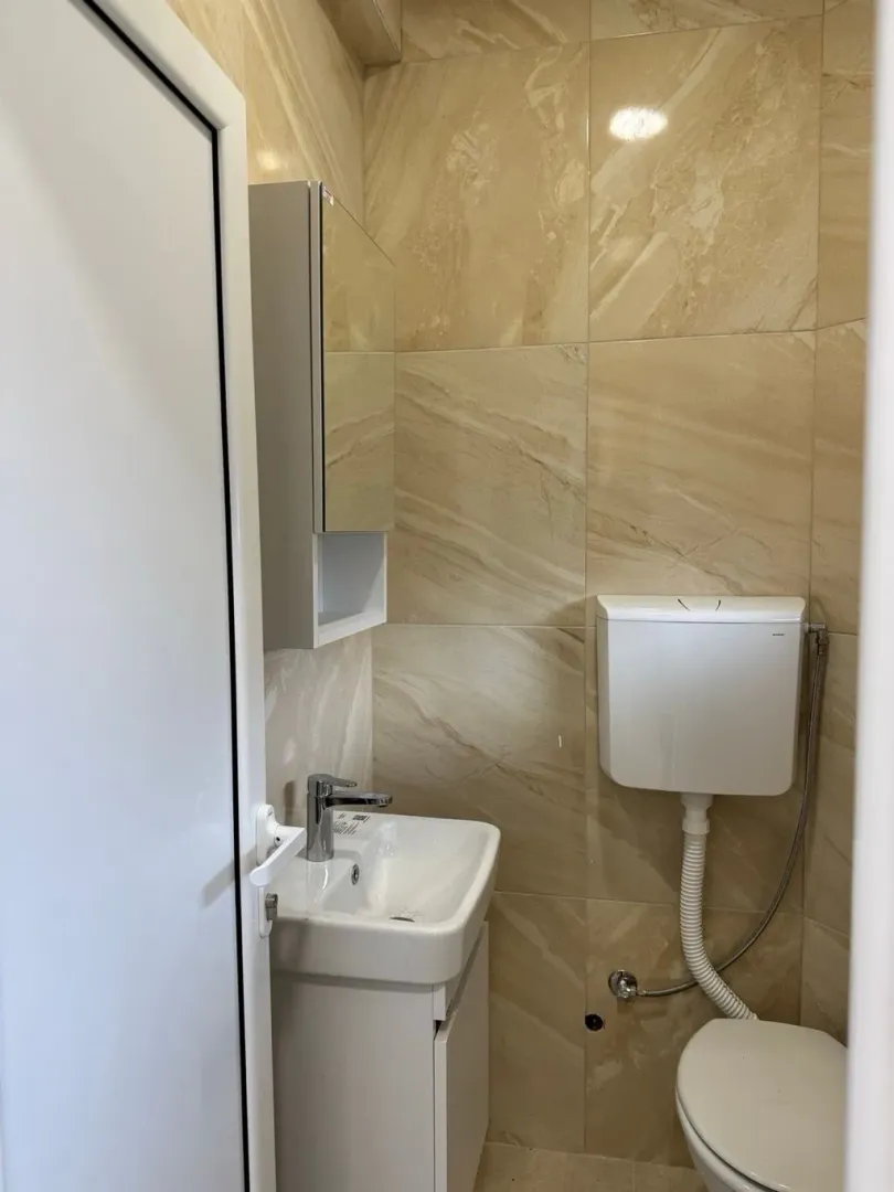 Manje kupatilo sa umivaonikom, ogledalom i wc šoljom obloženo keramičkim pločicama.