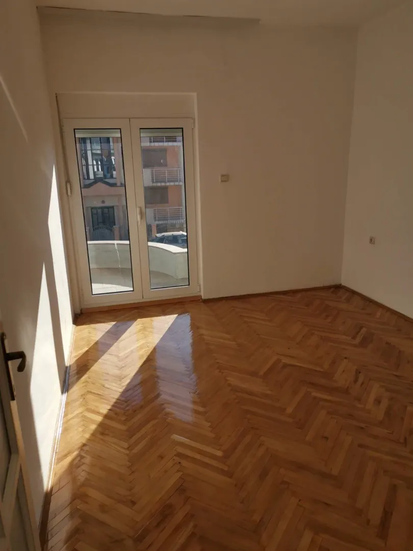 Prazna soba sa lakiranim parketom i balkonskim vratima.