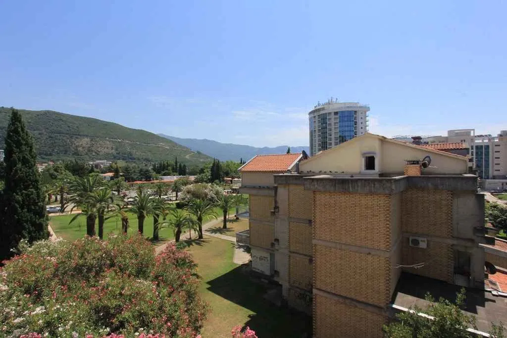 Pogled sa visine na uređeni park sa palmama, okolne zgrade i brda u daljini.