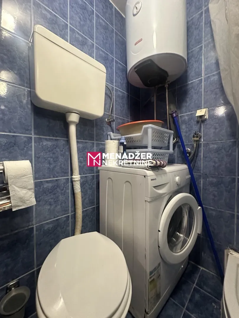 Manje kupatilo sa pločicama, wc šoljom, veš mašinom i bojlerom.