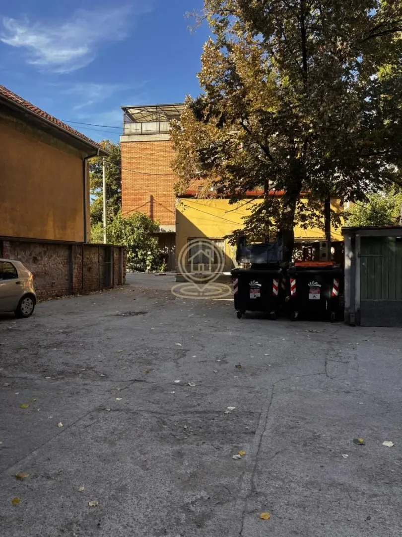 Asfaltirano dvorište sa okolnim zgradama i drvećem.