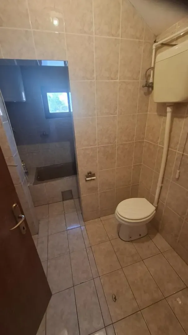 Manje kupatilo obloženo keramičkim pločicama sa WC šoljom i kadom u pozadini.
