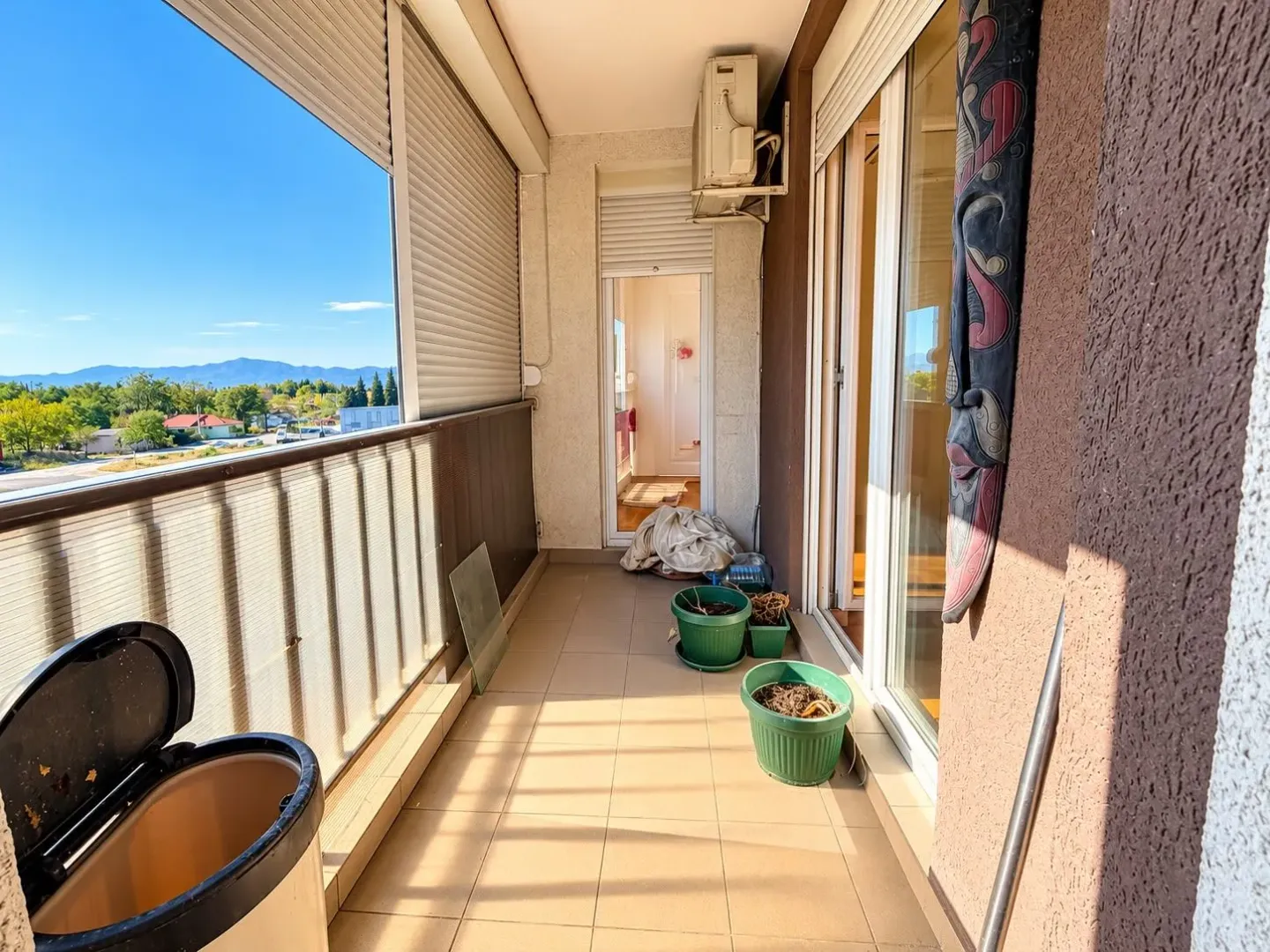 Dugački balkon sa pločicama, saksijama za cvijeće i pogledom na okolinu.