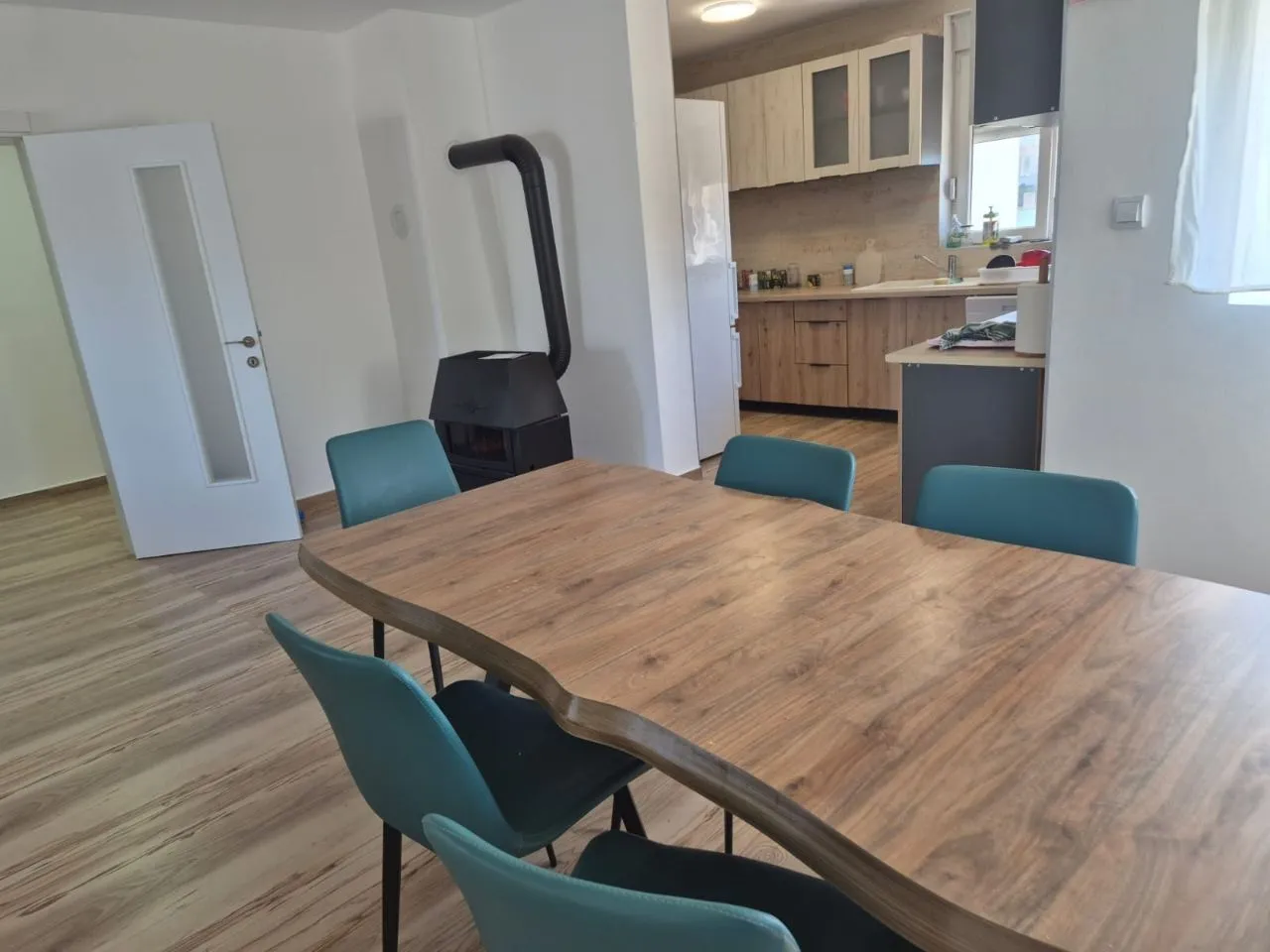 Dining area with a wooden table, blue chairs, and a wood-burning stove, with a view of the kitchen.