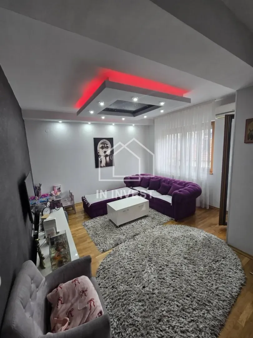 Dnevna soba sa ljubičastom garniturom, belim stočićem i spuštenim plafonom sa osvetljenjem.