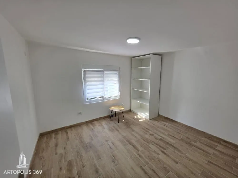 Empty room with laminate flooring, white walls, a window, a small table, and a white shelving unit.