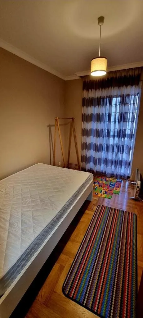 Manja spavaća soba sa krevetom, drvenim stalkom za odjeću i prozorom sa zavjesama.