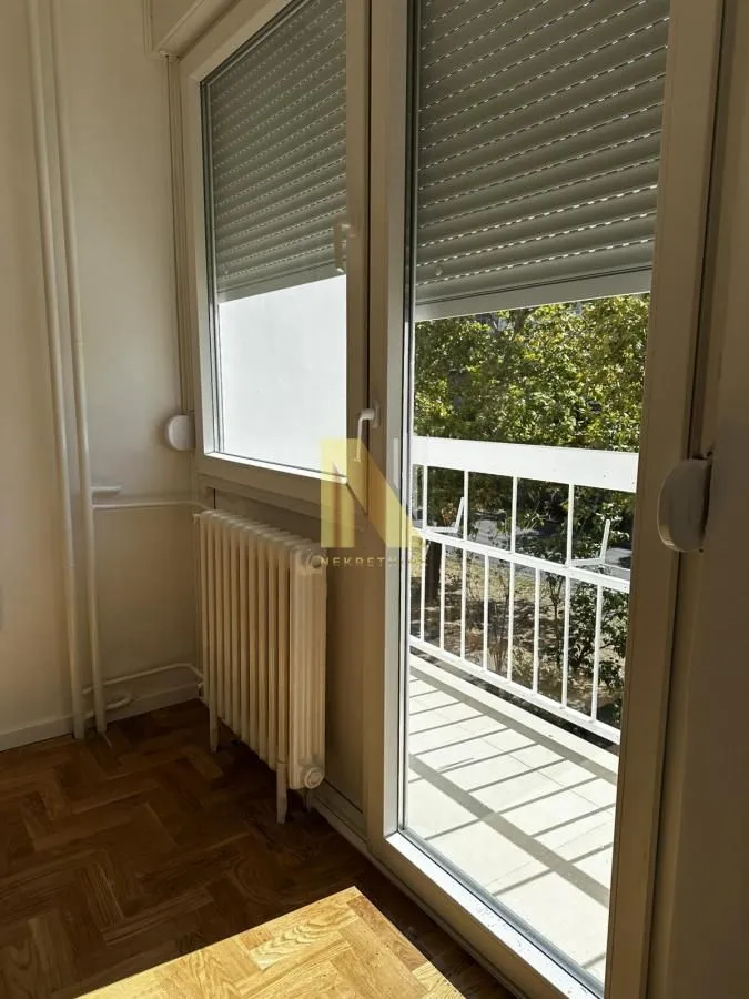 Pogled kroz balkonska vrata prema spoljašnjosti sa radijatorom i drvenim podom.